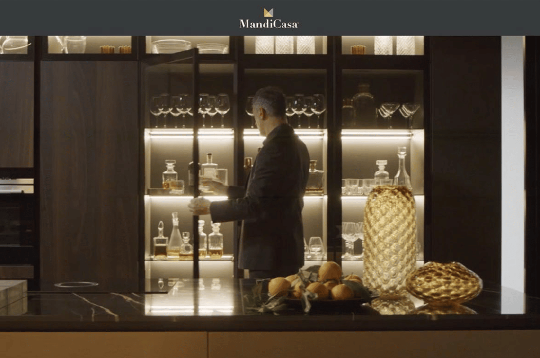 A man in a suit standing in a modern kitchen, reaching for a bottle of liquor from a glass-fronted cabinet.