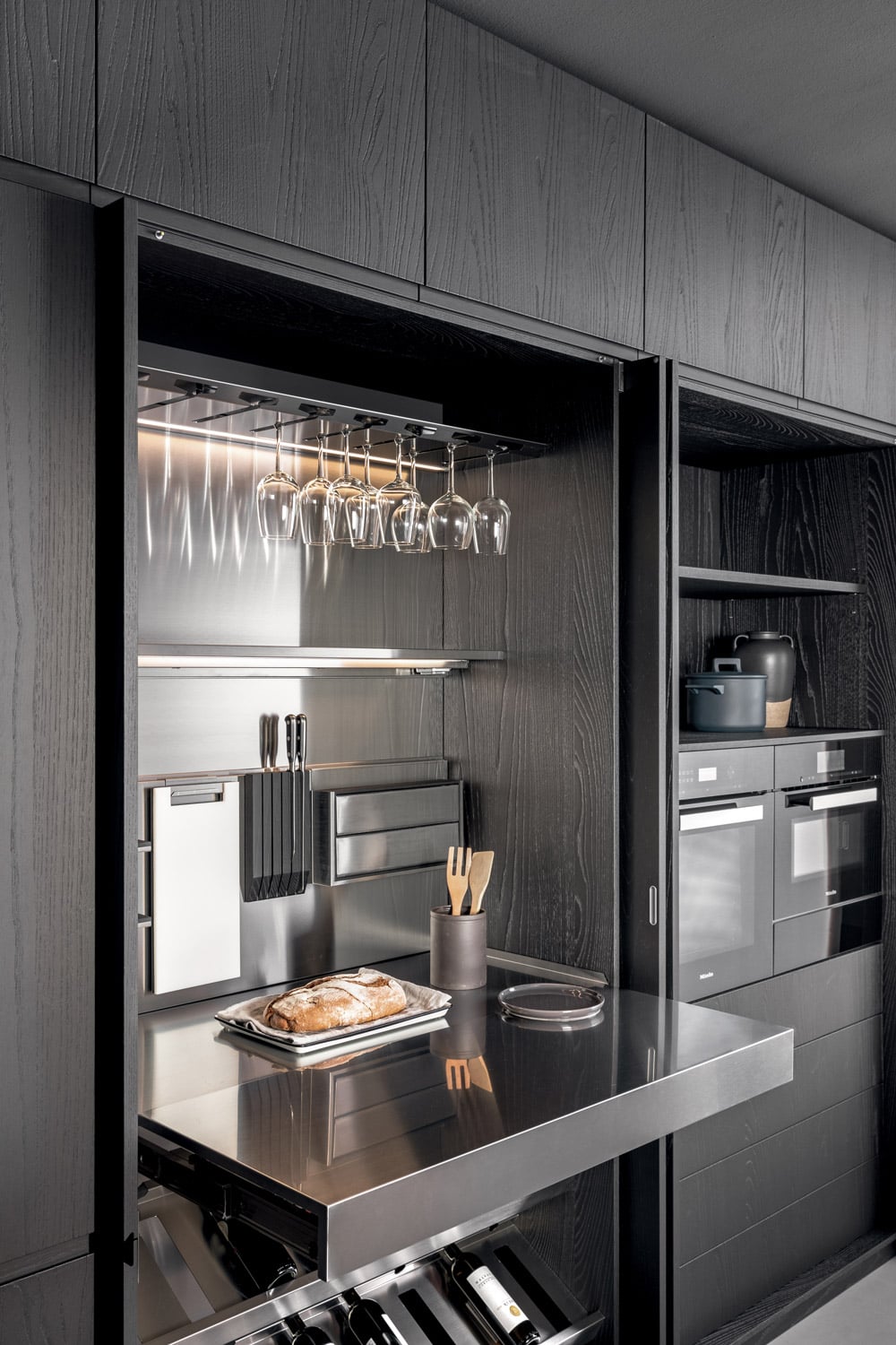Multifunctional tall unit customized to house wine racks, drawers, glassware, and knives while also including a full pull-out stainless steel worktop to extend the usable surface area.