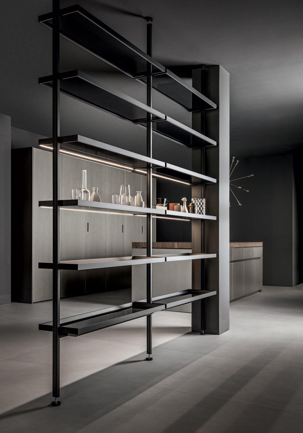To the side, the kitchen space bridges into the living area with a custom floor-to-ceiling open storage system featuring glass shelves with black lacquered metal profiles (same styling as the shelves inside the tall units). 