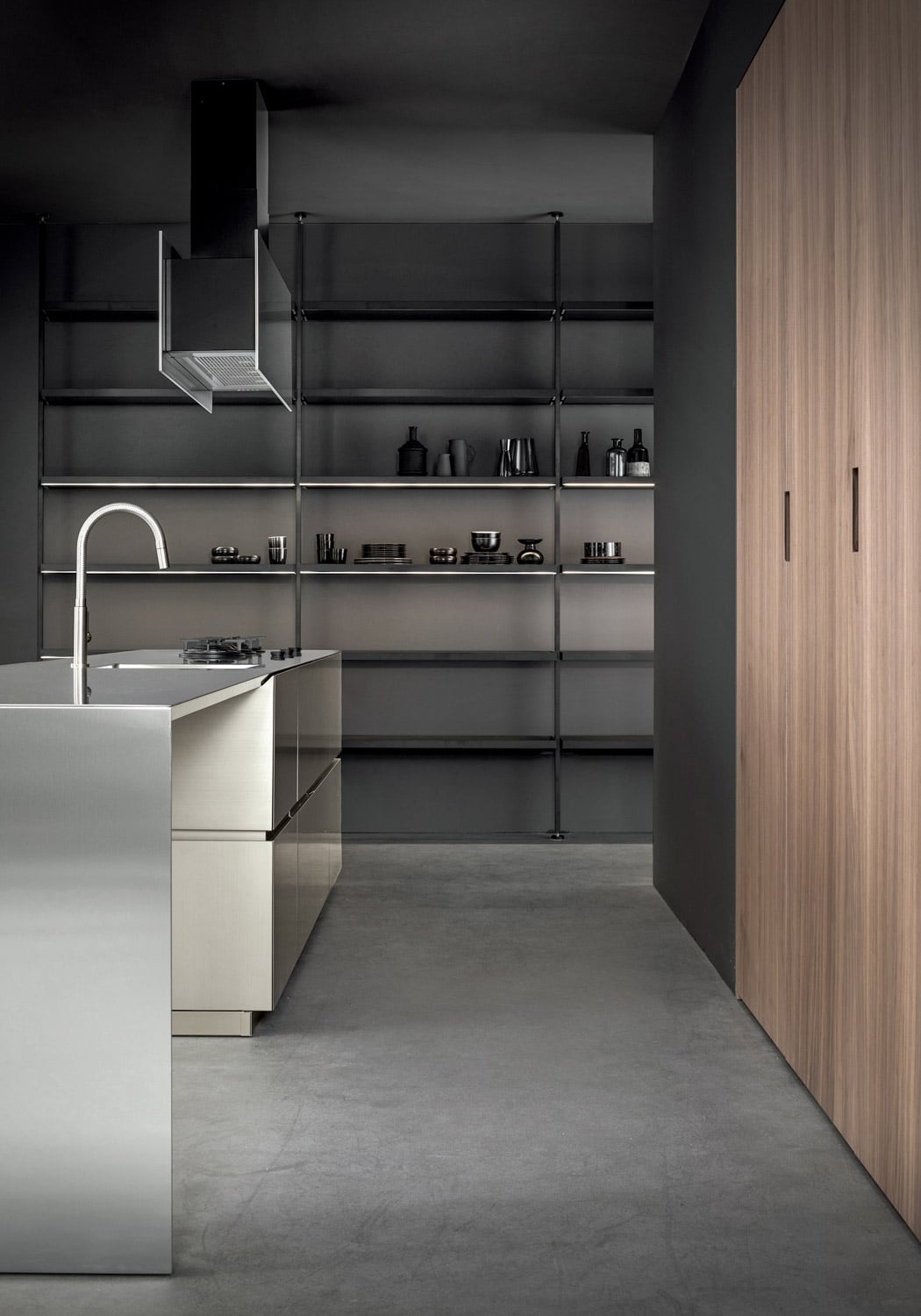 In the background, beyond the steel island, the wall is lined with a modern bookcase in black lacquered metal, with glass shelves and integrated led lights, bridging kitchen and living area.