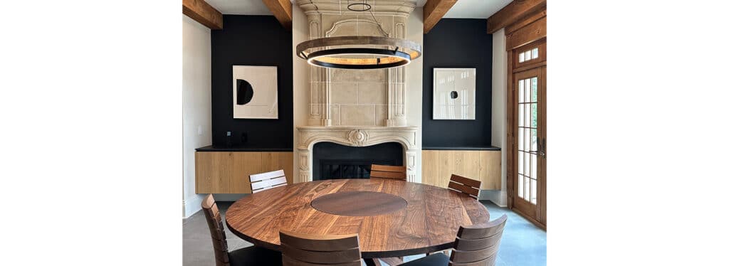 A modern dining room with a round wooden table, a stone fireplace, and a black wall with artwork.